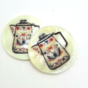 Puede incluir: Dos botones decorativos redondos y blancos con una imagen pintada de una cafetera vintage con diseños florales coloridos.