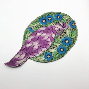 Op de afbeelding: Geborduurde patch met een paars en wit vogelontwerp. De vogel is geplaatst in een groene ovaal, versierd met blauwe bloemen en groene bladeren. Deze patch is waarschijnlijk bedoeld voor decoratief gebruik op kleding of tassen.