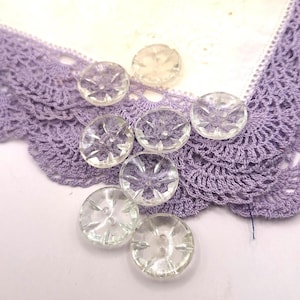 May include: Clear, faceted glass buttons with a starburst design are scattered on a white cloth with a lavender crocheted lace trim. The buttons have two holes for sewing and are approximately 0.75 inches in diameter.