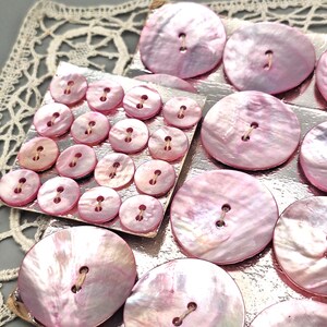 Könnte beinhalten: Eine Sammlung runder, irisierend rosa Knöpfe mit Perlmutt-Finish und zwei Löchern in der Mitte. Die Knöpfe variieren in der Größe und sind auf quadratischen, metallisch rosa Karten angeordnet, die eine glatte, reflektierende Oberfläche aufweisen.