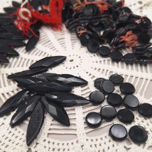 Könnte beinhalten: Eine Auswahl an schwarzen Vintage-Perlen und Verzierungen. Die Sammlung umfasst facettierte, blattförmige Perlen, runde Perlen und Perlen, die auf rotem und orangefarbenem Faden aufgefädelt sind. Die Perlen sind auf einer weißen Häkeldecke angeordnet.