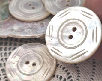 May include: A set of round, off-white mother-of-pearl buttons, each featuring two central holes and a series of engraved lines around the perimeter. The buttons have a smooth, iridescent surface with a subtle sheen and a circular recessed center.