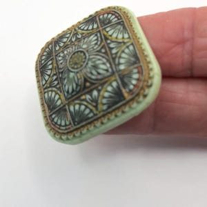 May include: A square, green button with a gold trim and a detailed, intricate design in black, gold, and white.