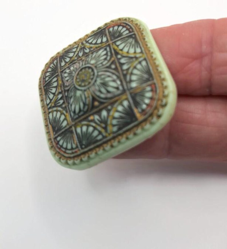 May include: A square, green button with a gold trim and a detailed, intricate design in black, gold, and white.