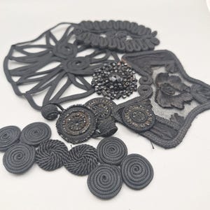 Könnte beinhalten: Eine Sammlung schwarzer Nähutensilien, bestehend aus mehreren runden Kordelknöpfen mit Spiralmustern, einer rechteckigen Spitzenapplikation mit Blumenstickerei, einem runden Perlenbesatz und einem großen dekorativen Kordelgeflecht.