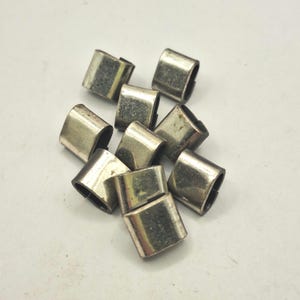 Pode incluir: Uma coleção de pequenos fechos de metal prateado. Cada fecho é retangular com bordas arredondadas e uma abertura curva. Os fechos estão espalhados sobre uma superfície branca, com alguns sobrepostos.