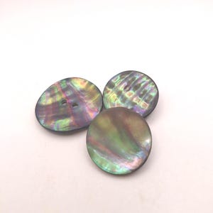 Może przedstawiać: Trzy iryzujące guziki z dwoma otworami każdy. Guziki mają perłowy połysk w kolorach zieleni, fioletu i złota. Guziki są okrągłe i owalne.