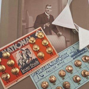 Puede incluir: Dos tarjetas de presentación de cartón vintage con botones de cuello de metal dorado. La tarjeta naranja muestra un caballo y un jinete con el texto The National Lever Studs. La tarjeta azul presenta una ilustración de un barco y el texto The Reliable Collar Studs.