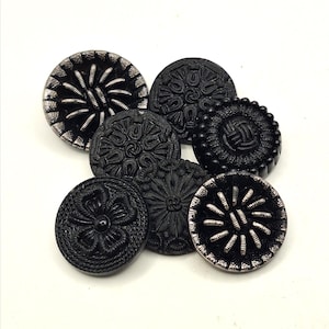 Può includere: Una collezione di sette bottoni neri con vari disegni in rilievo. Alcuni bottoni hanno bordi color argento, mentre altri presentano intricati motivi floreali o geometrici. I bottoni sono rotondi e di dimensioni diverse, adatti per cucito o artigianato.
