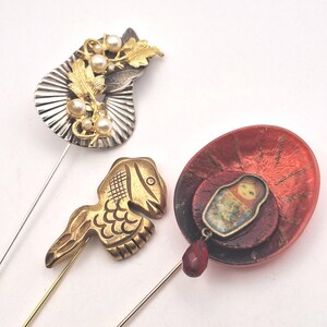 Può includere: Tre spille decorative per cappelli. Una spilla presenta un design a foglie argentate e dorate con perle finte. Un'altra è un pesce dorato. La terza spilla ha un design a strati rosso e marrone con un piccolo ciondolo a bambola russa e una perla a goccia rossa.