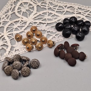 Può includere: Una collezione di bottoni vintage in vari stili e colori. Ci sono bottoni in metallo dorato, bottoni rotondi neri, bottoni rivestiti in tessuto marrone e bottoni fantasia. I bottoni sono disposti su una superficie grigio chiaro con un centrino di pizzo bianco.