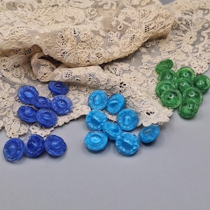 Op de afbeelding: Een verzameling ronde, doorschijnende knopen in tinten donkerblauw, lichtblauw en groen, elk met een centraal verhoogd cirkelvormig detail en een geschulpte rand, gerangschikt in drie afzonderlijke groepen op kleur op een oppervlak.