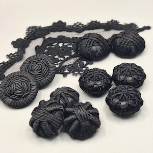 Pode incluir: Uma coleção de botões revestidos de tecido preto com padrões bordados intrincados e detalhes em relevo, acompanhados por um pedaço de renda preta com design floral, tudo disposto sobre uma superfície lisa de cor clara.