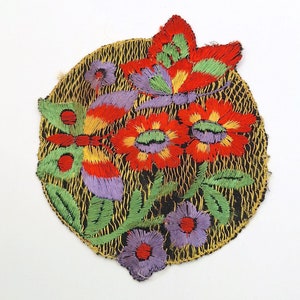 Op de afbeelding: Een ronde geborduurde patch met een bloemenontwerp met rode, gele en groene bloemen en twee vlinders. De achtergrond is een zwarte stof met een gouden gaasovertrek.