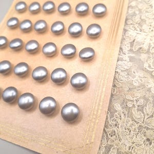 May include: A selection of silver-grey, pearlescent buttons displayed on a vintage card. The buttons are oval and have a glossy finish. Cream-coloured lace fabric is visible in the background, adding a touch of elegance to the display.