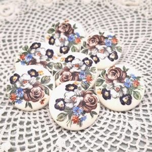 May include: Seven round, off-white ceramic buttons with floral designs. Each button features a bouquet of roses and other flowers in shades of brown, white, blue, and orange. The buttons are arranged on a white, lace-like surface.
