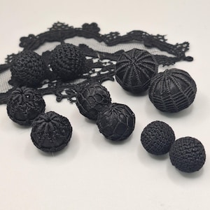 May include: A collection of black decorative buttons in various styles, including woven and textured designs. The buttons are arranged on a white surface with a piece of black lace in the background. These buttons are suitable for sewing and craft projects.
