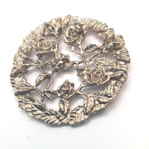 Pode incluir: Um pingente de metal prateado com um design circular com recortes intrincados de rosas e folhas.