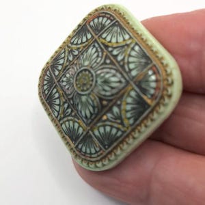 May include: A square-shaped button with a raised, intricate design. The button is made of a light green material and features a geometric pattern in shades of black, gold, and white.