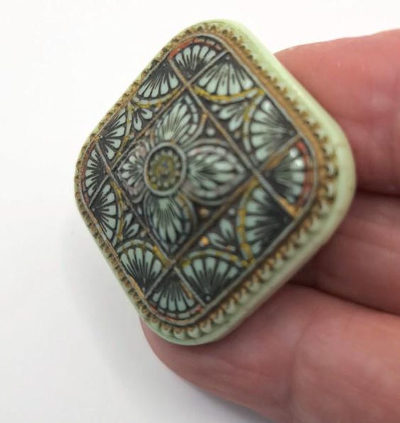 May include: A square-shaped button with a raised, intricate design. The button is made of a light green material and features a geometric pattern in shades of black, gold, and white.
