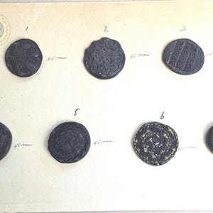 May include: Seven dark grey buttons with embossed patterns, including floral and spiral designs. Measurements in millimetres are noted next to each button. The buttons are displayed on a cream background.