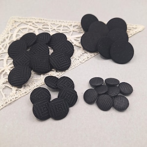 May include: A collection of black, round fabric-covered buttons. The buttons have a textured weave pattern and are arranged in several small groups. The background includes a white lace doily.