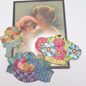 Op de afbeelding: Drie geborduurde stoffen patches met bloemen- en fruitmotieven. Eén patch toont een mand met oranje en gele bloemen, een andere een mand met roze en blauwe bloemen, en de derde een paarse mand gevuld met rood en geel fruit.