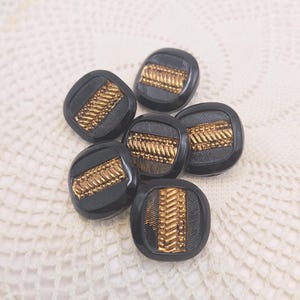 Puede incluir: Seis botones cuadrados negros con un diseño decorativo dorado. Los botones tienen un patrón texturizado en relieve en el centro. Los botones están dispuestos sobre una superficie blanca texturizada.