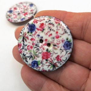 May include: Two round, white, wooden buttons with a floral print in shades of pink, blue, and green. The buttons have two holes.