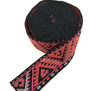 May include: A rolled-up black woven ribbon featuring a geometric pattern in red, pink, and brown. The ribbon's edge displays a detailed design. This decorative trim is suitable for craft projects. The ribbon is tightly wound.