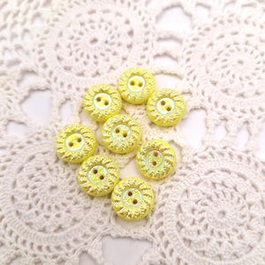 May include: Eight yellow buttons with a textured, sunburst design. Each button has two holes for sewing. The buttons are arranged on a white, lacy, crocheted doily. The buttons are approximately 2 cm in diameter.