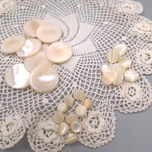 Puede incluir: Una colección de botones redondos de nácar en color blanco roto de varios tamaños, con una superficie lisa e iridiscente de brillo sutil. Los botones están dispuestos en tres grupos distintos, mostrando su lustre natural y su forma circular.