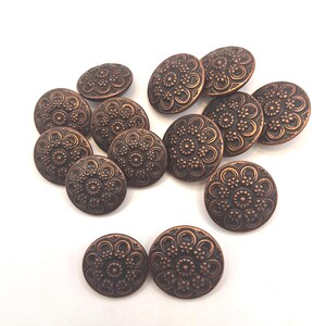 Puede incluir: Un conjunto de botones metálicos de color bronce con un diseño floral. Los botones son redondos y tienen un patrón detallado en relieve. Los botones están dispersos sobre una superficie blanca.