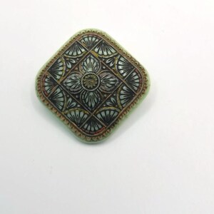 May include: A square-shaped, green ceramic brooch with a black and gold geometric design.