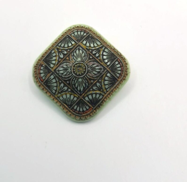 May include: A square-shaped, green ceramic brooch with a black and gold geometric design.