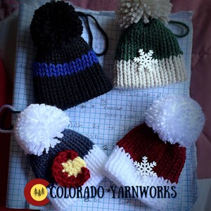 May include: Four crocheted hats with pom-poms. The hats are in various colors, including black, green, red, and blue. Each hat has a snowflake design on the front. The hats are on a sheet of graph paper with the text "COLORADO YARNWORKS" written on it.