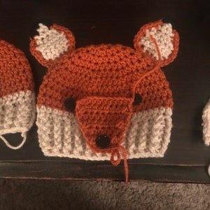 Puede incluir: Un gorro de punto de zorro hecho con hilo blanco y naranja. El gorro tiene dos ojos de botón negro y una nariz de botón negro.