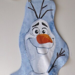 May include: A blue fabric stocking with a white snowman design. The snowman has a carrot nose, black eyes, and a black mouth. The snowman is wearing a white hat with a brown brim. The stocking is decorated with a snowflake pattern.
