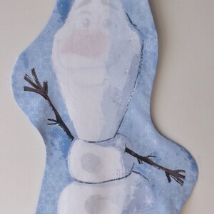 May include: A blue and white fabric wall hanging featuring a cartoon snowman with a stick figure body and a brown twig for arms. The background is a light blue with a snowflake pattern.