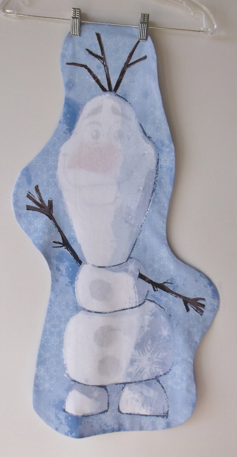 May include: A blue and white fabric wall hanging featuring a cartoon snowman with a stick figure body and a brown twig for arms. The background is a light blue with a snowflake pattern.