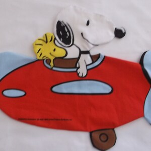 May include: Snoopy and Woodstock flying in a red airplane with blue accents. The airplane has a brown wheel. Snoopy is wearing a black helmet and a brown collar. Woodstock is yellow.
