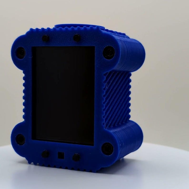 Puede incluir: Un objeto rectangular azul impreso en 3D con un panel frontal negro liso. El marco azul presenta un patr&oacute;n lateral texturizado y acanalado, con cuatro fijaciones circulares negras en las esquinas y detalles negros adicionales en los bordes.