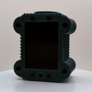 Puede incluir: Una funda protectora rectangular de color verde oscuro impresa en 3D con una superficie texturizada y granulada. La funda presenta una gran abertura rectangular central, esquinas redondeadas con hendiduras circulares.