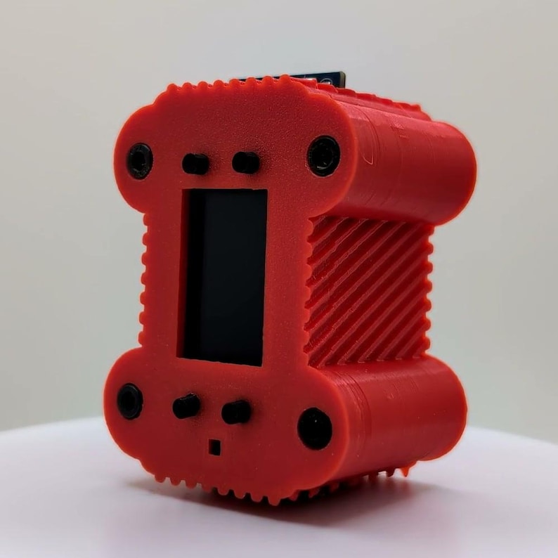 May include: A bright red, 3D-printed rectangular electronic housing with a central black display screen. The exterior features a textured, ribbed surface, four circular screw holes at the corners, and four small black protruding control buttons.