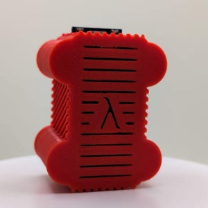May include: A bright red, spool-shaped object with a textured, ribbed surface. The center of the front face features a black Greek lambda symbol, surrounded by horizontal ventilation slits. The top edge shows a small black electronic component.