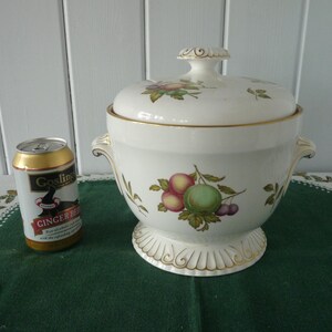 May include: A white ceramic lidded serving dish with a gold rim and handles, decorated with a fruit and floral design. A can of Gosling's Ginger Beer is next to the dish. The dish sits on a green cloth.