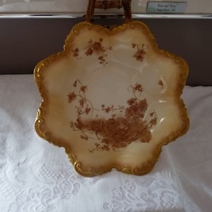 Könnte beinhalten: Eine große, weiße Keramikschale mit einem gewellten Rand und einem braunen Blumendesign. Die Schale hat einen goldenen Rand.