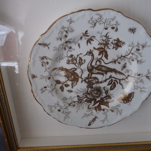 May include: A white porcelain plate with a gold rim and a gold design of birds and flowers. The plate is framed in a gold frame.