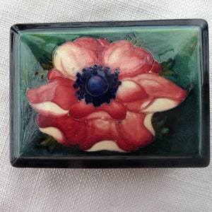 May include: A ceramic trinket box with a green background and a red and white flower with a blue center.