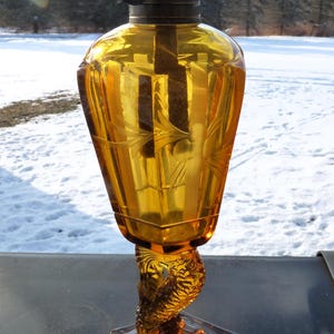 May include: Amber-colored antique oil lamp with a decorative etched glass design. The lamp features a detailed base and a metal top with a wick adjustment mechanism. The lamp's glass body has a unique shape and is set on a hexagonal base.